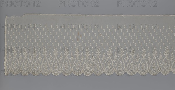 Lace fragment — shuttlecock; maker, mid-19th century, France. Lace for clothing accessories/fashion. Lower edge: semicircular serrations bordered with fine serrations with geometric ornament. Above the semicircular serrations: floral ornament repeating with a twig motif in triangular divisions; above this, a small twig in a regular projection. Material: lace. Width 42 cm, length 430 cm., A long horizontal piece of sheer fabric displayed against a gray background, featuring an overall delicate cream-colored embroidery; the fabric has a scalloped lower edge with intricate floral and vine motifs woven into a repeating ornate border, above which a pattern of small embroidered sprigs is scattered across the translucent field; the top edge is straight with some fraying and a few small holes and loose threads visible; the embroidery shows a slightly darker cream in places and there are a few small discolored spots on the fabric.