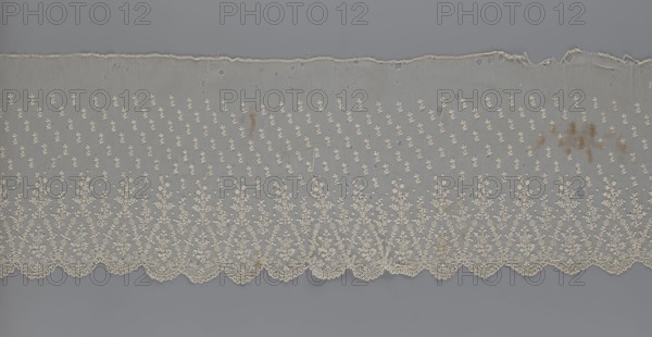 Lace fragment — shuttlecock; lace; clothing accessory; fashion. Lower edge: semicircular serrations bordered with fine serrations with geometric ornament. From the semicircular serrations upwards: a floral ornament repeating with a twig motif in triangular divisions; above this, a small twig in a regular projection. Mid-19th century, France. Material: lace. Width 42 cm, length 430 cm., A horizontal strip of delicate fabric with ornate embroidery lies against a flat background. The fabric is semi-transparent with a scalloped lower edge featuring dense floral and leafy embroidered motifs that form triangular arches along the border. Above the border, the textile has a repeating pattern of small floral sprigs scattered evenly across the field. The top edge is irregular and slightly frayed with several small holes visible throughout the upper portion of the fabric. There are scattered brownish stains on the right side and a few smaller discolorations elsewhere. The visible colors include o...