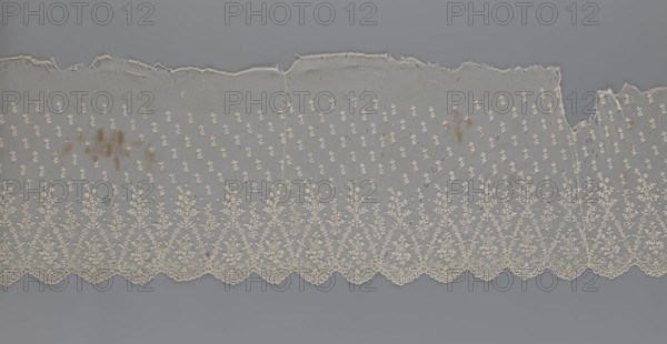 Lace fragment — shuttlecock; lace; clothing accessory; fashion. Lower edge: semicircular serrations bordered with fine serrations with geometric ornament. From the semicircular serrations upwards: a repeating floral ornament with a twig motif in triangular divisions. Above this, a small twig in a regular projection. Mid-19th century, France. Lace. Width 42 cm, length 430 cm., A long strip of delicate lace fabric laid flat against a gray background, showing a scalloped lower edge with an ornate floral embroidery pattern in off-white and cream tones, a repeated small floral motif across the body of the lace in similar off-white and cream, areas of frayed and uneven raw upper edge with some small holes, a few irregular darker brownish stains on the left and slightly toward the right, and several small irregular holes and worn spots scattered across the mesh.