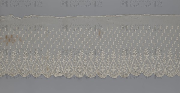Lace fragment — shuttlecock, mid-19th century, France. Lace shuttlecock for clothing accessory/fashion. Lower edge: semicircular serrations bordered with fine serrations with geometric ornament. From the semicircular serrations upwards: a repeating floral ornament with a twig motif in triangular divisions; above this, a small twig in regular projection. Width 42 cm, length 430 cm., A length of delicate lace fabric laid flat, showing a sheer net background in pale gray with an overall cream-colored embroidered floral motif; the embroidery forms a dense scalloped border along one edge with repeating vertical sprays of small cream flowers and scrollwork, and a sparser pattern of tiny cream floral clusters scattered above the border across the net; there are a few irregular holes and frayed areas near the opposite edge and several small brownish stains in places on the net; the background beneath the lace is a smooth medium gray.