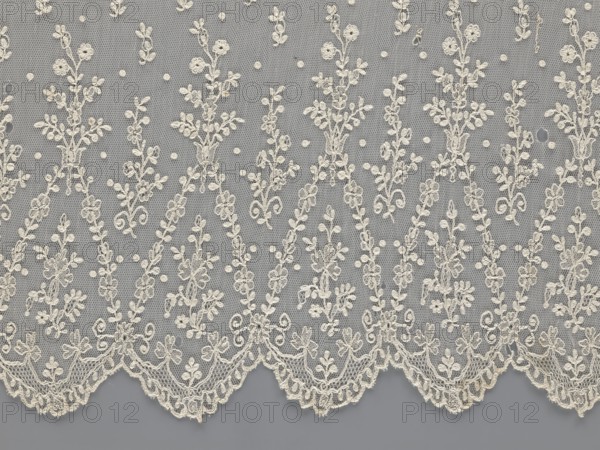 Lace fragment — shuttlecock; lace; clothing accessory; fashion. Lower edge: semicircular serrations bordered with fine serrations with geometric ornament. From the semicircular serrations upwards: a floral ornament repeating with a twig motif in triangular divisions. Above this, a small twig in a regular projection. Mid-19th century, France. Lace. Width 42 cm, length 430 cm., A delicate piece of lace fabric with an overall gray background and off-white or cream embroidered floral motifs, showing a repeating pattern of vertical floral sprays and small dot accents on a fine hexagonal mesh; the lower edge has a scalloped border with ornate scrollwork and floral clusters, and the embroidery includes leaves, tiny blossoms, and curling tendrils in the off-white or cream color against the gray.