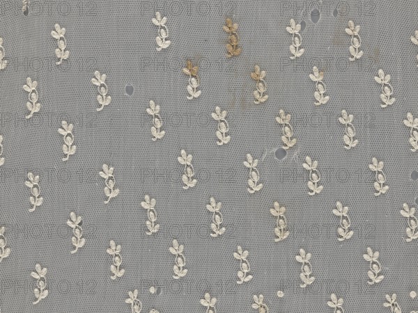 Lace fragment — shuttlecock;, Mid-19th century, France. Lace clothing accessory fashion. Lower edge: semicircular serrations bordered with fine serrations with geometric ornament. From the semicircular serrations upwards: a floral ornament repeating with a twig motif in triangular divisions. Above this, a small twig in a regular projection. Material: lace. Width 42 cm; length 430 cm., A sheer net fabric with a repeating floral motif embroidered in off-white thread over a gray background, showing a few small holes and a group of light brown stains near the top, with the netting forming a hexagonal mesh pattern throughout.