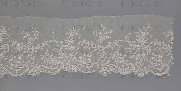 Lace fragment — shuttlecock; maker, purchase provenance; fashion, clothing accessory; needle lace and bobbin lace shuttlecocks; tiny spiders — needlework and bobbin ornament elements on factory-made tulle; floral ornament with a large repeat; serrated lower edge with stylized feathery leaves and flower calyces, above them floral bouquets and flower projections; 1st half of the 19th century, France; lace; width 39 cm, length 328 cm, A length of delicate lace fabric laid flat against a smooth background, showing an ornate floral and scrollwork embroidered border with scalloped lower edge; the lace is primarily off-white or cream with intricate motifs of flowers, leaves, vines, and looping paisley-like shapes, set on a fine translucent netting that appears pale gray where visible, and the background around the lace is a uniform medium gray.