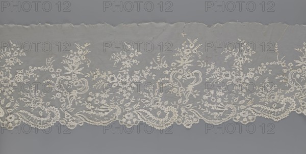 Lace fragment — shuttlecock;, needle lace and bobbin lace; shuttlecocks; tiny spiders — needlework and bobbin ornament elements on factory-made tulle. Floral ornament with a large repeat. On the serrated lower edge are stylized feathery leaves and flower calyces; above them are floral bouquets and flower projections. 1st half of the 19th century, France. Lace; width 39 cm; length 328 cm., The image shows a horizontal piece of delicate lace fabric against a gray background, featuring an ornate floral pattern with a scalloped lower edge. The lace is off-white or cream in color, with intricate embroidered flowers, leaves, and vine motifs spread across a fine netted ground. The top edge of the fabric appears torn or uneven, and there are subtle darker spots or stains visible on the netting. The scalloped border includes circular and looped decorative elements, and the overall texture appears fine and airy with visible embroidery density variations. The predominant colors visible are cream,...