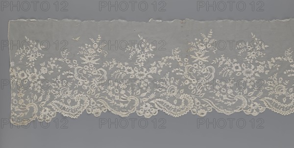 Lace fragment — shuttlecock;, Fashion accessory: needle lace and bobbin lace shuttlecocks; tiny spiders—needlework and bobbin ornament elements on factory-made tulle. Floral ornament with a large repeat. Serrated lower edge with stylized feathery leaves and flower calyces; above them are floral bouquets and flower projections. 1st half of the 19th century, France. Lace; width 39 cm, length 328 cm., A wide horizontal piece of delicate lace fabric laid flat, showing an intricate floral embroidered pattern along one long edge; the lace is primarily off-white or cream with fine threadwork forming stylized flowers, leaves, scrolls, and scalloped edging, set against a muted gray background that contrasts with the pale lace, and the fabric shows slight irregularities along the top edge and small areas of darker discoloration.