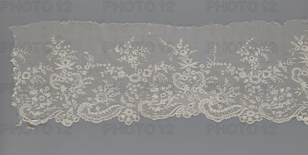 Lace fragment - shuttlecock;, Fashion accessory: clothing lace combining needle lace and bobbin lace—shuttlecocks, tiny spiders as needlework and bobbin ornament elements on factory-made tulle. Floral ornament with a large repeat. Serrated lower edge with stylized feathery leaves and flower calyces; above them floral bouquets and flower projections. 1st half of the 19th century, France. Lace; width 39 cm, length 328 cm., A wide length of delicate lace fabric laid flat, showing an intricate floral and foliate embroidery pattern on a fine net ground; the lace features a scalloped lower edge with ornate scrolls and stylized flower motifs and a straighter, slightly frayed upper edge; the embroidered motifs include roses, buds, leaves, and tendrils arranged in repeating clusters and garlands, with some areas displaying denser embroidery and others more open net; visible colors are shades of off-white and cream for the lace against a neutral gray background.