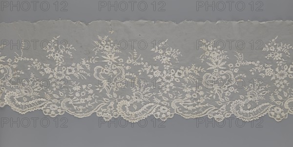 Lace fragment — shuttlecock., Purchase provenance. Fashion; clothing accessories. Lace: needle lace and bobbin lace; shuttlecocks; tiny spiders — needlework and bobbin ornament elements on factory-made tulle. Floral ornament with a large repeat. Serrated lower edge with stylized feathery leaves and flower calyces; above them are floral bouquets and flower projections. 1st half of the 19th century, France. Lace, width 39 cm, length 328 cm., A horizontal strip of delicate lace with a scalloped lower edge, displayed against a flat background; the lace features an intricate floral pattern with bouquets, leafy sprigs, and scrolling vine motifs woven throughout, and there are some small irregular holes and tiny stains visible on the fabric; the lace appears in off-white and cream tones, set against a muted gray backdrop with slightly darker gray at the top and bottom edges.