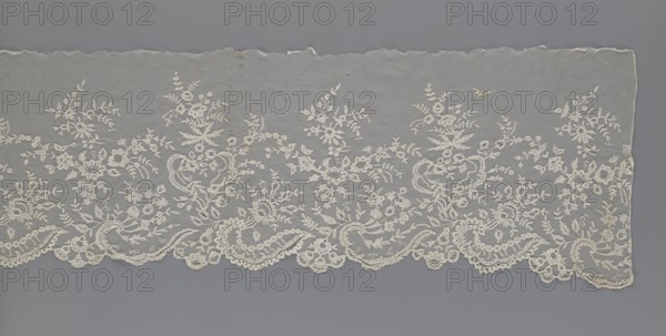 Lace fragment — shuttlecock;, Purchase provenance. Fashion; clothing accessories. Lace needle lace, bobbin lace; shuttlecocks; tiny spiders — needlework and bobbin ornament elements on factory-made tulle. Floral ornament with a large repeat. Serrated lower edge with stylized feathery leaves and flower calyces; above them are floral bouquets and flower projections. 1st half of the 19th century, France. Lace, width 39 cm, length 328 cm., A length of delicate lace fabric laid flat, showing an intricate floral and foliate embroidery pattern in off-white on a muted gray background; the lace edge is scalloped with ornate looping motifs, and the upper edge appears slightly uneven; visible colors include off-white, cream, and various shades of gray.