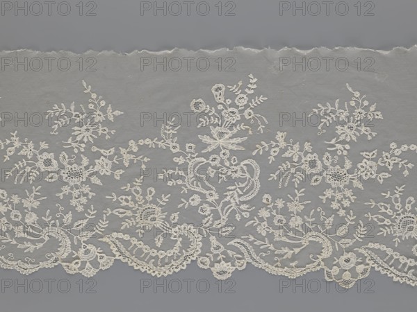 Lace fragment — shuttlecock; creator, purchase provenance; fashion; clothing accessories; lace; needle lace; bobbin lace; shuttlecocks; tiny spiders — needlework and bobbin ornament elements on factory-made tulle; floral ornament with a large repeat; serrated lower edge with stylized feathery leaves and flower calyces; above them floral bouquets and flower projections; 1st half of the 19th century; France; lace; width 39 cm; length 328 cm, A length of delicate lace fabric with an ornate floral and foliate pattern laid flat against a plain background; the lace shows a scalloped lower edge with curved motifs and a few openwork areas, and the upper edge appears slightly torn or uneven. The design features a group of embroidered flowers, leaves, tendrils, and small circular elements arranged in a repeating, asymmetrical layout, with sections of net-like mesh between the motifs. The visible colors are shades of off-white for the lace and a muted gray for the background.