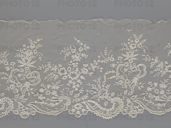 Lace fragment - shuttlecock., Purchase provenance. Fashion; clothing accessory. Needle lace and bobbin lace; shuttlecocks; tiny spiders—needlework and bobbin ornament elements on factory-made tulle. Floral ornament with a large repeat. Serrated lower edge with stylized feathery leaves and flower calyces; above them are floral bouquets and flower projections. 1st half of the 19th century, France. Lace. Width 39 cm; length 328 cm., A piece of delicate lace fabric pictured against a neutral background, showing an intricate floral and scrollwork embroidery in a creamy off-white color on a muted gray ground; the lace edge is scalloped with a few torn and frayed areas along the top, and the design includes a group of stylized flowers, leaves, and looping vine motifs distributed across the fabric with varied openwork and solid embroidered textures in shades of off-white and cream set against the gray backdrop.
