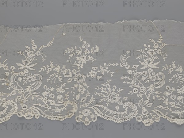 Lace fragment — shuttlecock;, Purchased provenance. Fashion accessory; needle lace and bobbin lace; shuttlecocks. Tiny spiders — needlework and bobbin ornament elements on factory-made tulle. Floral ornament with a large repeat. Serrated lower edge with stylized feathery leaves and flower calyces; above them are floral bouquets and flower projections. 1st half of the 19th century, France. Lace; width 39 cm, length 328 cm., A length of delicate lace fabric in shades of white and off-white lies against a gray background; the lace features an intricate floral and foliate pattern with a scalloped lower edge, showing clusters of stylized flowers, leaves, tendrils, and ornamental loops arranged in a repeating, flowing design, with some areas of slightly darker off-white thread and subtle shadowing that emphasize texture and raised embroidery.