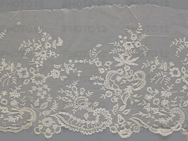 Lace fragment — shuttlecock, purchase provenance; fashion; clothing accessories. Needle lace and bobbin lace with shuttlecocks; tiny spiders — needlework and bobbin ornament elements on factory-made tulle. Floral ornament with a large repeat. Serrated lower edge with stylized feathery leaves and flower calyces; above them floral bouquets and flower projections. 1st half of the 19th century, France. Lace; width 39 cm, length 328 cm., A delicate off-white lace panel lies flat against a muted gray background, showing an intricate floral and foliate pattern worked in fine thread. The lace features a scalloped lower edge with ornate curved motifs and a few decorative circular medallions, while the field above contains a group of stylized flowers, leaves, and tendrils arranged in flowing sprays and connected by slender stems. Several areas display denser embroidered clusters contrasting with open net-like sections, and there are some tiny imperfections and loose threads along the top edge. T...