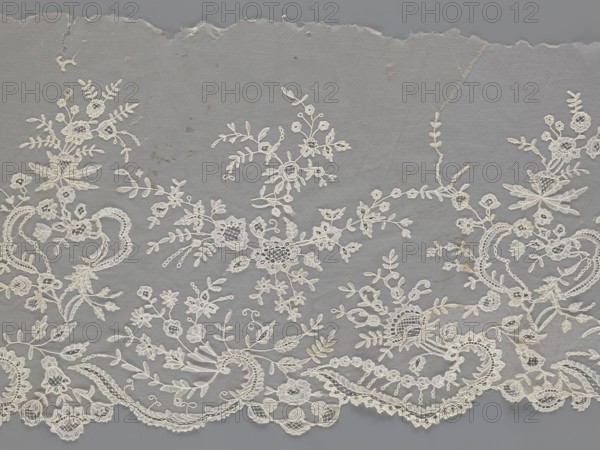 Lace fragment - shuttlecock;, Fashion accessory: clothing ornament. Materials/technique: needle lace and bobbin lace with tiny spider motifs—needlework and bobbin ornament elements on factory-made tulle. Design: shuttlecocks; floral ornament with a large repeat; serrated lower edge with stylized feathery leaves and flower calyces; above them floral bouquets and flower projections. Date: 1st half of the 19th century. Place: France. Object type: lace. Dimensions: width 39 cm, length 328 cm., A delicate piece of lace with an off-white floral embroidery laid against a gray background, showing a scalloped lower edge and an irregular torn upper edge; the lace features a variety of floral and foliate motifs including blossoms, leaves, and tendrils arranged in a flowing, repeating pattern, with areas of denser embroidered clusters and more open netting, and small blemishes or darker specks scattered on the fabric.