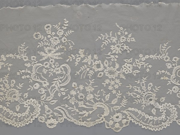 Lace fragment - shuttlecock;, purchase, Fashion accessory; clothing ornament. Materials/technique: needle lace and bobbin lace, shuttlecocks; tiny spiders—needlework and bobbin ornament elements on factory-made tulle. Design: floral ornament with a large repeat; serrated lower edge with stylized feathery leaves and flower calyces; above them floral bouquets and flower projections. Date: 1st half of the 19th century. Place: France. Object type: lace. Dimensions: width 39 cm, length 328 cm., A length of delicate fabric with an overall muted gray background and an intricate off-white lace overlay, showing a repeating floral and foliate pattern. The lace includes several stylized flowers, leaves, and curling tendrils rendered in off-white thread, with areas of open mesh and denser embroidered motifs. Along the lower edge there is a scalloped border of ornate lacework with curved ornamental shapes and small circular motifs. The top edge appears slightly irregular with a torn or frayed finis...