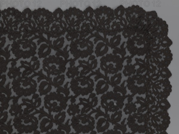 Cape, lace, in the form of a square with two wide, elongated lace ties. Continuous floral ornament with a motif of a large, asymmetrical flower sprig. Raised edge with a strip of floral edge ornament made separately and sewn on. 2nd half of the 19th century, France. Height 97 cm, width 168 cm., A close-up of a piece of black lace fabric with an ornate floral and leaf pattern set against a gray background, showing a scalloped edge and dense embroidered motifs in black and dark gray tones.