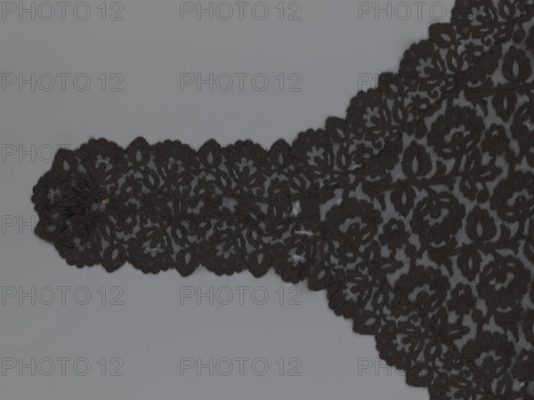 Cape, lace, in the form of a square with two wide, elongated lace ties. Continuous floral ornament with a motif of a large, asymmetrical flower sprig. Raised edge; a strip of floral edge ornament made separately and sewn on. 2nd half of the 19th century, France. Height 97 cm, width 168 cm., A section of black lace fabric with a floral and leaf pattern lies against a smooth grey background, showing scalloped edges and a denser floral motif on one side that tapers into a narrower strip with repeating flower shapes, all rendered in black against the grey surface.