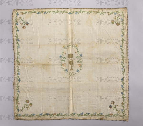 Church napkin. Napkin made of linen fabric with a rare plain weave. Embroidered with chain stitch using colored silk and gold thread also pierced. In the center, in a wreath of flowers tied with ribbon, is a gold chalice, and above it a gold oval—probably a patera, not a host. In the corners are floral sprigs tied with a ribbon with double loops. A floral tendril runs around the edges. The flowers are stylized rather than purely decorative. Flower colors: green, brown, blue, sky blue, pink, tobacco. Narrow gold lace sewn around the edges now very damaged. Fabric density: 30 weft threads, 23 warp threads per cm. 2nd half of the 18th century, Poland. Materials: linen, silk, gold thread. Height 100 cm, width 105 cm., A square light beige fabric with visible creases and faint stains has a decorative embroidered border of green vine-like stems with small blue and brown flowers and leaves; the corners show clusters of similar flower motifs in brown, blue, and green. The center contains an em...