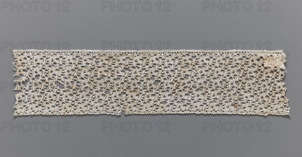 Lace fragment, bobbin lace, one edge finished with picots, 17th century, Flanders; width 6.5 cm, length 31 cm., No background. Repossession, A rectangular piece of openwork lace laid flat against a smooth background, showing an intricate repeating pattern of interconnected loops and small circular openings; the lace appears off-white to beige with subtle darker brownish spots or stains in places, and the background is a solid medium gray; edges of the lace are slightly irregular with a few frayed threads and tiny loose ends, and the lace surface shows slight variations in tone and texture across its length.