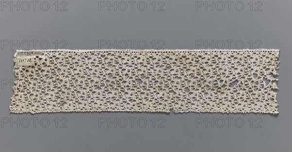 Lace fragment, bobbin lace, one edge finished with picots. 17th century, Flanders. Width 6.5 cm, length 31 cm., No background. Repossession, A rectangular strip of crocheted lace in off-white and beige tones lies flat against a smooth gray background, showing an openwork pattern of interconnected loops and small circular holes; the top and bottom edges run mostly straight with slight irregularities while the left and right ends are frayed, and a small rectangular fabric tag is attached near the top left corner; there are a few tiny darker brown spots on the lace and subtle variations in the off-white color across the piece.