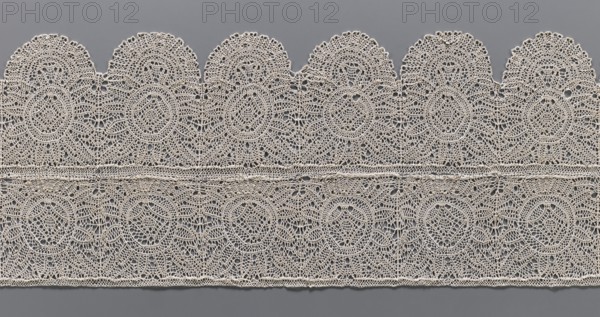 Lace fragment, bobbin lace, 17th century, Flanders. Two laces sewn together across their entire width: one with straight edges, the other with strongly semicircularly serrated edges. Both feature identical rosette motifs, with the serrated lace’s rosettes framed at the bottom by a fan-shaped motif. Lace; width 21.5 cm, length 71 cm., A horizontal strip of intricately patterned lace in an off-white or cream color lies against a flat gray background, showing a repeating scalloped top edge and a straight bottom edge, with a few horizontal bands separating two tiers of openwork designs; the lace features several circular and oval motifs made from fine interlacing threads, creating a delicate mesh of loops and small openings, with scallop-shaped arches along the top and a uniform border along the bottom, and the overall visual texture is detailed and lacy in cream contrasted with the smooth medium-gray background.
