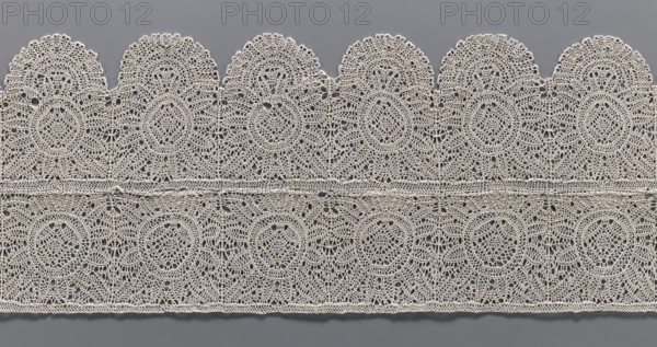Lace fragment, bobbin lace, two pieces sewn together across their width: one with straight edges, the other with strongly semicircularly serrated edges. Both feature identical rosette motifs; in the serrated piece the rosettes are framed at the bottom by a fan-shaped motif. 17th century, Flanders. Lace; width 21.5 cm, length 71 cm., A horizontal strip of delicate lace displayed against a gray background, showing a repeating scalloped edge at the top and a straight finished edge at the bottom, with an intricate openwork pattern of circular motifs and interlaced loops; the lace appears in a light off-white or cream color contrasted with the medium gray behind it, and the textile shows fine thread work, small holes, and symmetrical decorative elements throughout.