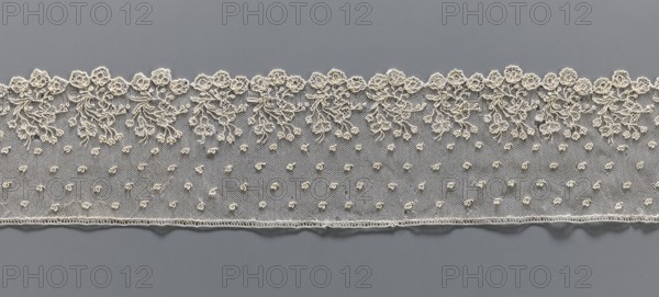 Lace fragment, repossession provenance; needle lace, Alençon lace, 18th century, France. The edge features a broad floral ornament composed of a repeated, elaborate bouquet of flowers; the edge is serrated following the forms of the flower calyxes, repeated in threes. Above, against the mesh background, very small heart-shaped leaves are depicted. Lace; width 8 cm, length 51 cm., A horizontal strip of delicate lace in off-white is laid against a smooth gray background; the lace features a scalloped top edge with an ornate floral and leafy embroidery pattern and a lower edge finished with a fine stitched border, while the central area of the lace is a fine net dotted with a few small raised embroidered buds.