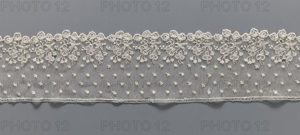 Lace fragment, needle lace Alençon style, 18th century, France. The edge features a broad floral ornament of repeated elaborate bouquets; the edge is serrated following the forms of flower calyxes repeated in threes. Above, against the mesh background, very small heart-shaped leaves are depicted. Lace fragment, width 8 cm, length 51 cm., A horizontal strip of delicate lace sits against a smooth background, showing an upper scalloped edge with ornate floral embroidery and flowing vine shapes, and a lower straight edge with a subtle hem; the lace field contains a repeating dotted textured pattern. The visible colors include off-white and cream in the lace contrasted with a muted gray background.