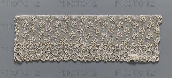 Lace fragment, needle lace, Alençon lace, one-sided geometric edge ornament with alternating ovals and quatrefoils; above, against a mesh background, asymmetrical twigs and dots are projected. 18th century, France. Lace; width 9 cm, length 29 cm., A rectangular piece of delicate lace displayed against a gray background, showing a fine hexagonal net ground in a beige or off-white color with a scalloped lower edge, an ornate lower border of interconnected oval and floral loop motifs in beige with raised corded outlines, and a field above scattered with a few small looped floral sprigs and dots in the same beige tone; the overall palette is beige and off-white set on gray.