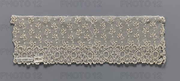 Lace fragment, needle lace, Alençon lace, one-sided geometric edge ornament including alternating ovals and quatrefoils; above, against a mesh background, asymmetrical twigs and dots are projected. 18th century, France. Width 9 cm, length 29 cm., Repossession, A rectangular piece of delicate lace displayed flat against a gray background, showing a fine hexagonal net ground with a pattern of small clustered floral motifs and raised dots across the field; the lower edge features a decorative scalloped border of interconnected oval and circular motifs with complex looping outlines and openwork, while the top edge is straighter and more irregular; the lace is an off-white or creamy color contrasting with the medium gray background.