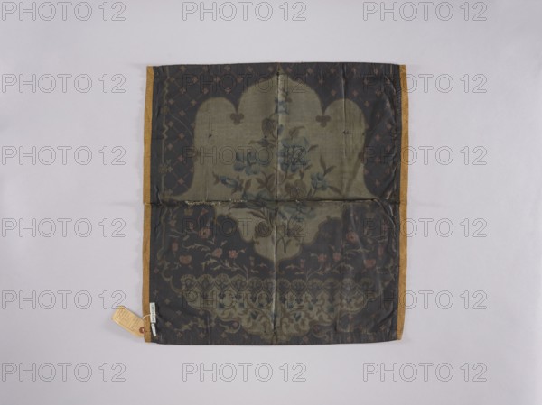 Tablecloth, church paraments; printed fabric with flowers, leaves, and plant ornaments; sacred art for churches and chapels furnishings; 19th century; place of origin, fabric; height 63.5 cm, width 59.8 cm, A square piece of fabric with a dark central panel and a patterned border in a muted gold color, laid flat on a pale gray background; the central area shows a faded floral motif in shades of olive green, blue, dark brown, and muted pink, with a darker overlay creating scalloped shapes and lattice-like accents; the fabric has visible fold lines and a few small dark spots, and a small tag attached near one corner shows a bit of beige and red but its markings are not described.
