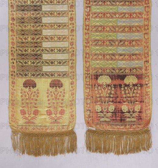Kontusz belt, Bipartite heads with a motif of elongated flower bushes of various species with carnations at the top, alternating transverse stripes with a plant twig and a motif of elongated rosettes with rhombuses. Floral twig border. Sewn-in silk fringe, yellow, braided at the top. Colors: straw, brown, white, green, and blue. At the bottom of one of the heads a lying FS preserved. 2nd half of the 18th century, Kraków, national costume, clothing accessory. Length 396 cm, width 38 cm., Two long narrow woven textile panels hang side by side, each with a decorative border and fringe at the bottom. The left panel has a pale yellow ground with horizontal patterned bands near the top in darker tones and a floral motif in coral and brown below, framed by a coral floral border and finished with a gold fringe. The right panel has a warmer, more reddish background with greenish and bluish horizontal patterned bands and a darker rectangular area near the lower portion featuring cream floral mot...