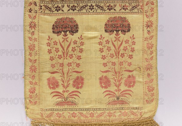 Kontusz belt, Bipartite heads with a motif of elongated flower bushes of various species with carnations at the top, alternating transverse stripes with a plant twig and a motif of elongated rosettes with rhombuses; floral twig border. Sewn-in yellow silk fringe, braided at the top. Colors: straw, brown, white, green, and blue. At the bottom of one of the heads, a preserved lying FS signature. 2nd half of the 18th century; Kraków place of origin?. National costume clothing accessory. Length 396 cm, width 38 cm., A rectangular fabric panel with a pale yellow background featuring a symmetrical floral pattern in reddish-pink and dark brown, showing a pair of tall stylized flowering stems with clustered blossoms and leafy branches, framed by a narrow decorative floral border along the sides and top, and a wider floral band along the bottom; the main field has subtle creases and a small area of wear, and the bottom edge is finished with a woven fringe in a golden-beige tone.