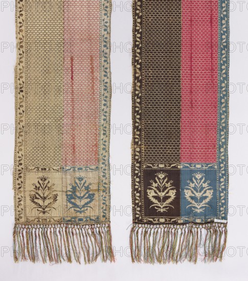 Kontusz belt, Bipartite heads with flower bush motifs in carp scale; edge with a motif of small flower sprigs arranged in a tendril. Colorful silk fringe, sewn on. Colors: gold, brown, silver, and blue. Signed at the bottom of the heads: FS lying. 2nd half 18th century. Slutsk, Grand Duchy of Lithuania; Poland. National costume, clothing accessory. Length 356.5 cm; width 28.5 cm., Two long woven textile panels hang side by side against a pale background; each panel has a patterned field with a scallop-like repeating motif and a decorative border with a floral vine. The left panel shows a muted beige ground transitioning into a faded pink vertical stripe with a narrow blue floral edge and a beige outer border, and its lower section includes a stylized floral medallion in darker brown and blue tones. The right panel displays a darker brown vertical field next to a bright pink stripe and a blue border, with a lower panel containing a contrasting floral motif in light beige on brown and bl...
