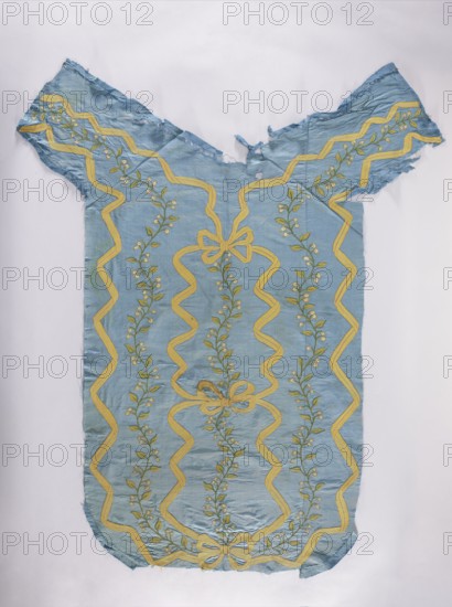 Back of a chasuble made of blue silk rep, embroidered with ribbon motifs tied in the center with three bows and delicate plant tendrils; motifs include flowers, leaves, and plant ornaments. Predominant colors: blue background, yellow, green, and white. Liturgical vestment, sacred art; churches and chapels furnishings. 4th quarter of the 18th century, Poland. Length 123 cm, width 67 cm., A single piece of fabric shaped like a loose garment is displayed on a plain background; the fabric is primarily light blue with a repeating decorative pattern in yellow and green, featuring sinuous yellow ribbon-like lines forming large wavy borders and bow shapes, and green vine motifs with small leaf shapes running between the yellow lines; the fabric shows frayed and uneven edges and a few small areas of wear or thinning, and the overall surface has subtle folds and creases.