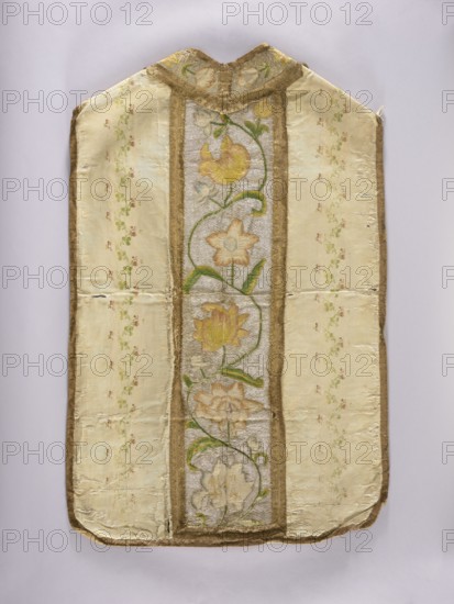 Chasuble, liturgical vestment, plant ornaments, church paraments, sacred art, churches and chapels furnishings. The column is embroidered with colorful silks with large flowers on a silver background. The sides are made of silk grosgrain brocaded with silk thread and motifs of a winding ribbon stripe and branches with small flowers and clover leaves. The columns are embroidered on a background laid with silver thread with zigzag motifs, wide winding branches with various flowers. The neckline is similarly embroidered. The edges of the columns and neckline are edged with wide gold braid. The edges of the chasuble are trimmed with narrow braid. 18th century, Poland, length 107 cm, width 69 cm back, A long, narrow textile with a rounded top and bottom displayed against a pale gray background, showing a central vertical panel with a floral vine motif flanked by wider panels with a subtle scattered floral pattern; the central panel features stylized flowers and leaves in shades of yellow, p...