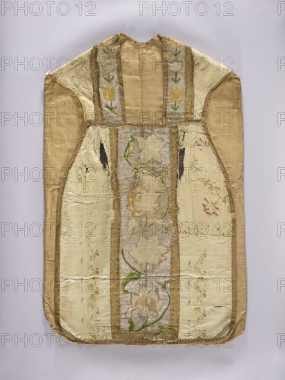 Chasuble, gift, liturgical vestment with plant ornaments, church paraments, sacred art, churches and chapels furnishings. The column is embroidered with colorful silks with large flowers on a silver background. The sides are made of silk grosgrain brocaded with silk thread and motifs of a winding ribbon stripe and branches with small flowers and clover leaves. The columns are embroidered on a background laid with silver thread with zigzag motifs, wide winding branches with various flowers. The neckline is similarly embroidered. The edges of the columns and neckline are edged with wide gold braid. The edges of the chasuble are trimmed with narrow braid. 18th century, Poland; length 107 cm, width 69 cm back., A flat garment-like textile laid against a pale neutral background, showing a central vertical panel with floral embroidery framed by narrow gold trim, flanked by lighter panels of patterned fabric and wider gold-colored side sections; the embroidered flowers and vines appear in sha...