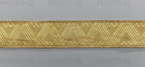 Gallon, origin unknown, galloons haberdashery, geometric ornaments, smaller snake pattern at the edges, early 19th century, Europe, fabrics, width 4.7 cm, length 53 cm, A horizontal woven band with a repeating geometric pattern featuring triangular and chevron motifs in shades of gold and pale yellow, set against a neutral gray background; the band shows textured stitching and subtle variations of golden tones.