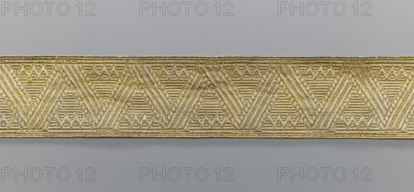 Gallon, galloons haberdashery, provenance unknown, geometric ornaments, smaller snake pattern at the edges, early 19th century, Europe, fabrics, width 4.9 cm, length 52 cm, A horizontal woven band with a repeating geometric pattern runs across the center of the image against a smooth background; the band displays triangular and chevron shapes formed by textured raised threads, and the visible colors include shades of gold, beige, cream, and light gray, with the background showing muted gray tones.