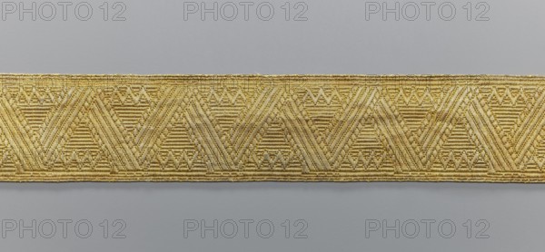 Gallon, galloons haberdashery, provenance unknown, geometric ornaments, a smaller snake pattern at the edges, early 19th century, Europe, fabrics, width 4.9 cm, length 52 cm, A horizontal woven trim with a repeating geometric pattern runs across the image, showing a group of chevron and triangular motifs formed by parallel textured lines; the trim appears gold with variations of lighter and darker gold tones against a neutral gray background.