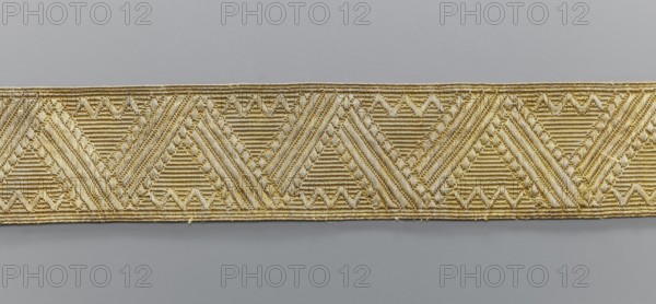 Gallon, galloons haberdashery, origin unknown, geometric ornaments, smaller snake pattern at the edges, early 19th century, Europe, fabrics, width 5.1 cm, length 41.8 cm, A narrow woven band with a repeating geometric pattern showing a row of connected triangular motifs formed by alternating diagonal and horizontal ribbed textures; the band appears in shades of gold and beige against a smooth light gray background.