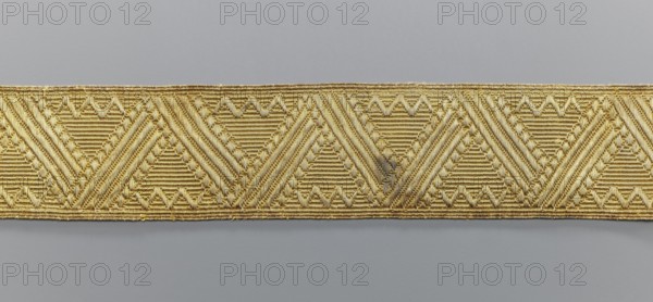 Gallon, galloons haberdashery, origin unknown, geometric ornaments, smaller snake pattern at the edges, early 19th century, Europe, fabrics, width 5.1 cm, length 41.8 cm, A narrow decorative band runs horizontally across the image, featuring a repeating geometric pattern of triangular motifs and diagonal ribbed textures. The band is primarily gold in color with variations of lighter and darker golden tones, and it has a slightly shiny, metallic sheen. The background above and below the band is a smooth, muted gray with subtle tonal variation. The gold band shows some areas with faint darker discoloration and small irregularities in the threads, and the edges of the band are slightly uneven with tiny loose fibers.