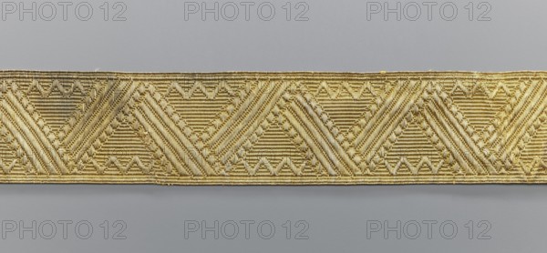 Gallon, galloons haberdashery, origin unknown, geometric ornaments, smaller snake pattern at the edges, early 19th century, Europe, fabrics, width 5 cm, length 53.6 cm, A horizontal band of woven material stretches across the image against a plain background, showing a repeating geometric pattern of triangular and chevron shapes created by tightly packed threads. The band has a metallic sheen and appears in shades of gold and light yellow, with highlights and subtle variations in tone where the weave changes direction. The background above and below the band is a smooth gray, slightly darker at the bottom and lighter toward the top. The woven surface displays ridged textures and diagonal lines formed by the thread, with small raised loops and interlaced details that create a tactile, embossed appearance.