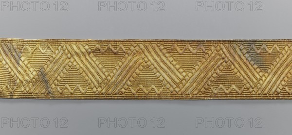 Gallon, galloons haberdashery, origin unknown, geometric ornaments, smaller snake pattern at the edges, early 19th century, Europe, fabrics, width 5 cm, length 53.6 cm, A horizontal strip of woven fabric with a repeating geometric pattern dominated by triangular and diagonal motifs in metallic gold tones sits against a neutral gray background; the fabric shows variations of bright gold and darker antique gold with small areas of muted grayish discoloration, and the woven texture reveals parallel ridges and raised braided details throughout the strip.