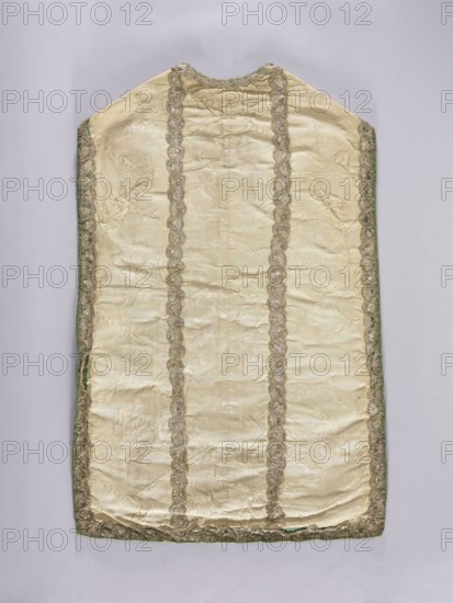 Chasuble, liturgical vestment, 18th century, possibly France. Against a background of broken lines, wavy-winding sprigs of large flowers and plant ornaments are arranged in branches. Division into columns is emphasized by stitching of silver lace, supplemented in places with a different type. Remnants of decorative stitching of green glass beads and sequins. Lining made of thin green silk. Function: church parament/sacred art. Dimensions: length 103 cm; width 68 cm back. Report dimensions: width 27.5 cm; height 36–38 cm., A long, sleeveless textile panel laid flat, predominantly cream colored with a subtle satin sheen and faint woven floral motifs; it has a scalloped metallic trim running along the outer edges and forming two parallel vertical bands and a band near the top, the trim appearing in a silver-gold tone. A narrow green edging is visible along the sides and bottom, and the fabric shows gentle wrinkles and slight creasing. The background is a pale gray surface. Colors visible ...