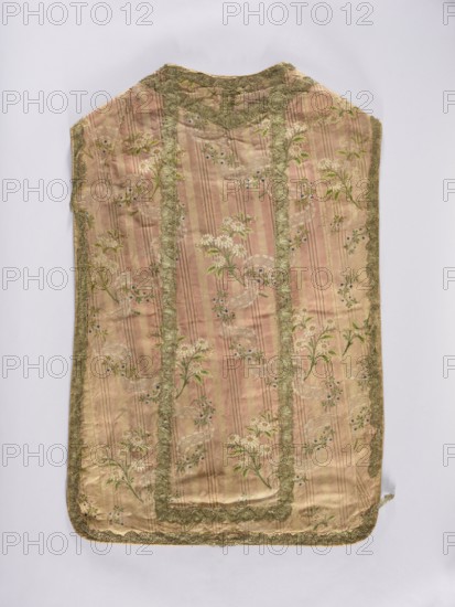 Chasuble, liturgical vestment; large floral pattern and lace motif set against a raspberry background. Dominant colors: brown, silver, and green. Entire piece trimmed with period lace; divided into columns with sewn-on gold lace. 2nd half of the 18th century, Lyon, France. Length 99 cm, width 66 cm back., A rectangular garment laid flat with rounded corners and a narrower top, showing a striped silk fabric in pink and beige tones patterned with floral sprigs in green and white and small blue accents, edged and paneled with a decorative metallic lace in a muted gold-green color, with visible pleats and subtle sheen on the fabric and some slight wrinkles and worn areas.