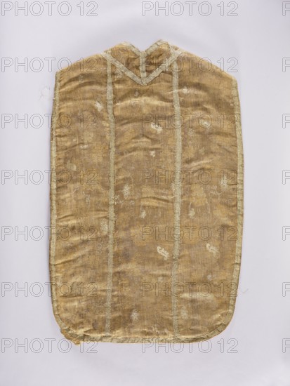 Chasuble, liturgical vestment, liturgical vestments, flowers, plant ornaments, church paraments, sacred art, churches and chapels furnishings. Divided into columns with sewn-on silver braid; edges trimmed with the same braid. 18th century, France. Length 101 cm, width 63 cm back., A single elongated garment-like textile displayed flat against a pale background, with a rounded bottom edge and a V-shaped notch at the top; the fabric appears worn and slightly wrinkled, showing a muted golden-brown base with lighter golden and pale beige woven decorative motifs and vertical bands; narrow pale trim runs along the outer edges and down the length in a few parallel lines, and the surface shows subtle variations in sheen and texture with some small areas of wear and fraying.