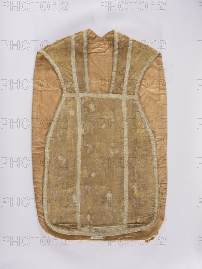 Chasuble, liturgical vestment, 18th century, France. Division into columns with sewn-on silver braid; edges trimmed with the same braid. Features flowers and plant ornaments; used as church paraments/sacred art for churches and chapels. Length 101 cm, width 63 cm back., An elongated fabric garment laid flat, with a rounded lower edge and a narrow upper neck opening, showing a central panel bordered by vertical and horizontal bands; the fabric appears worn and has a textured brocade pattern visible in places. The visible colors include shades of beige, tan, gold, and light cream, with the central panel slightly darker and more golden than the surrounding tan fabric. The bordering bands are a lighter cream color with frayed edges, and there are areas of subtle sheen and irregular wrinkles across the material. The garment edges show some wear and loose threads, and the surface displays small patched or faded spots where the pattern is less distinct.