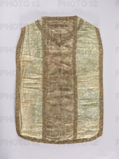 Chasuble, gift; silk liturgical vestment, 18th century, France. Church paraments with floral and leaf plant ornaments. Made of patterned silk fabrics: sides are striped two-tone damask with blue floral motifs; composed of columns of two types of fabric. Front column: brown, gray, and white silk with a pattern of small floral sprigs, report height 5.5 cm, report width 4.5 cm. Back column: silk of the same colors with a fine pattern of leaves arranged in an alternating rhythm sometimes left, sometimes right, report height 13.5 cm. Edges and columns trimmed with gold and silver lace. Function: liturgical vestment. Dimensions: length 98 cm; width 69 cm back. Some panel widths and report widths unspecified., A long sleeveless garment laid flat with a central vertical panel flanked by two lighter side panels, all made of a patterned woven fabric; the central panel is a darker muted gold with floral motifs and the side panels are a pale greenish-gold with similar floral patterns, the whole pi...
