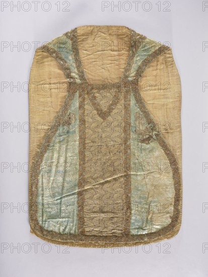 Chasuble, silk liturgical vestment, 18th century, France. Chasuble made of patterned silk fabrics: sides of stripe two-tone damask with floral motifs in blue; columns of two types of fabric — front column of brown, gray, and white silk with a pattern of small floral sprigs; back column of silk in the same colors with a fine pattern of leaves arranged in an alternating rhythm sometimes left, sometimes right. Edges and columns trimmed with gold and silver lace. Subject motifs: flowers, leaves, plant ornaments; function: church parament / liturgical vestment. Dimensions: length 98 cm; width 69 cm back. Fabric report heights and widths: sides — height of the repeat 31 cm; front column — height of the repeat 5.5 cm, width of the repeat 4.5 cm; back column — height of the repeat 13.5 cm., A garment laid flat with a rounded top and hem, showing a central panel and tapered side panels; the fabric appears silk-like with a textured, slightly wrinkled surface. The main colors visible are gold, pa...