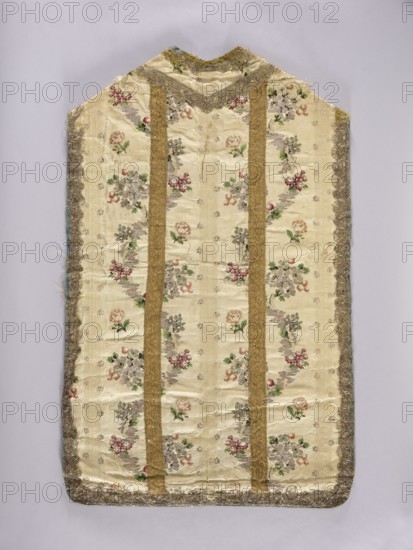 Chasuble, floral ornaments with flowers and leaves, columns on the back highlighted with wide gold braid; blue linen lining. Liturgical vestment liturgical parament, sacred art; churches and chapels furnishings. 18th century, Lyon. Length 107 cm, width 67 cm back., A single sleeveless garment laid flat, with a pale cream background patterned with floral motifs in colors including pink, green, grey, and muted red, and with a few scattered small decorative accents; two vertical bands in a gold tone run down the front, and the edges are trimmed with a metallic lace in gold and silver hues; the garment shows areas of slight wrinkling and wear, and the surrounding background is a soft light grey.